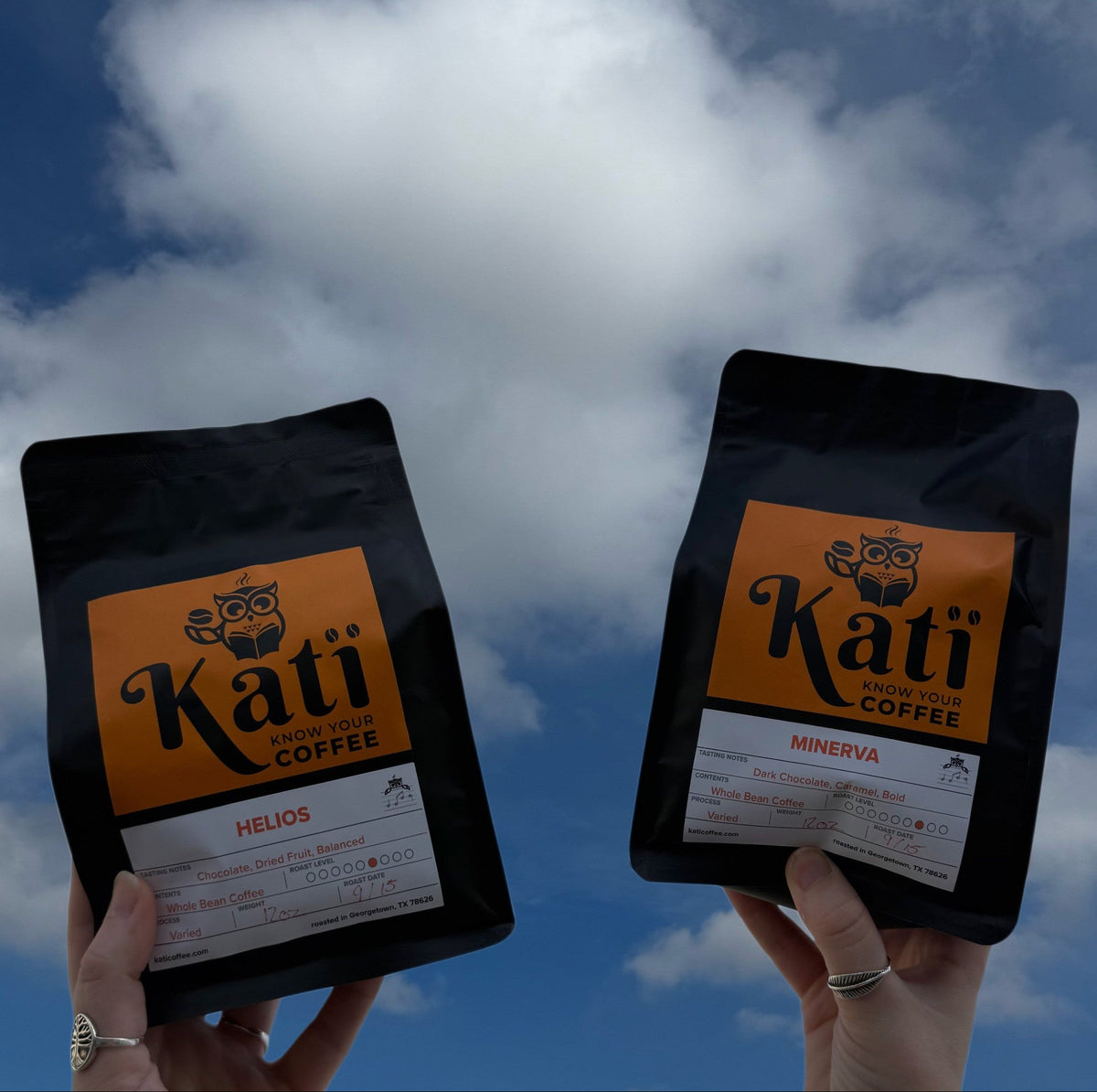 Two hands holding black Kati Coffee bags against a blue sky with white clouds. The left bag is labeled HELIOS and the right bag is labeled MINERVA
